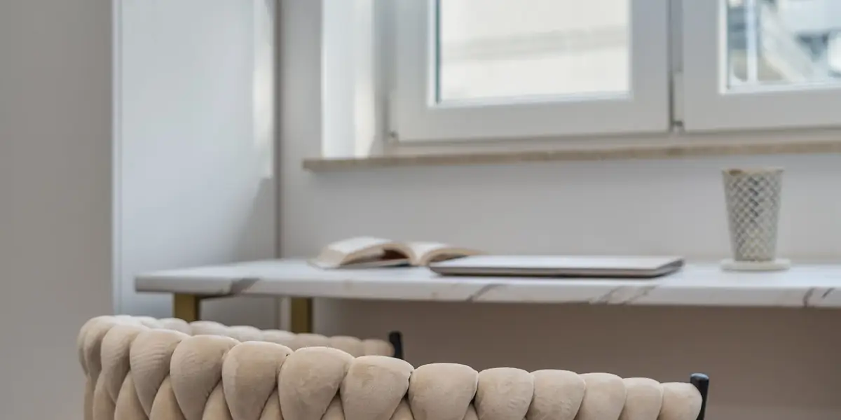 Upholstered woven chair at a marble-top desk beneath a window in a bright home workspace