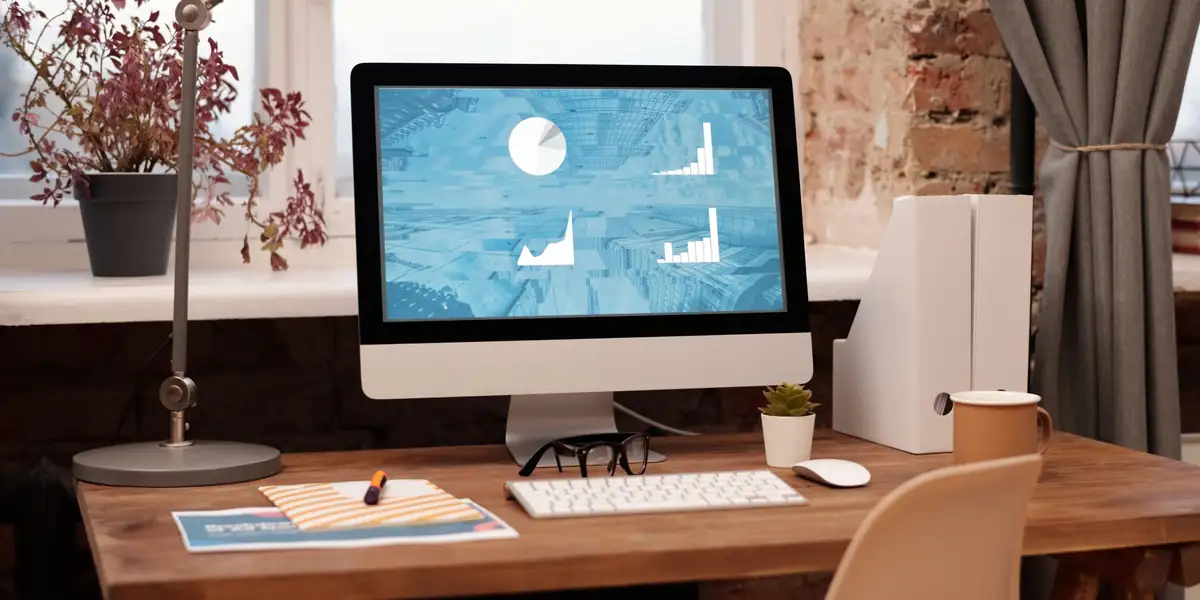 Wooden desk with a desktop computer, desk lamp and plants beside a bright window
