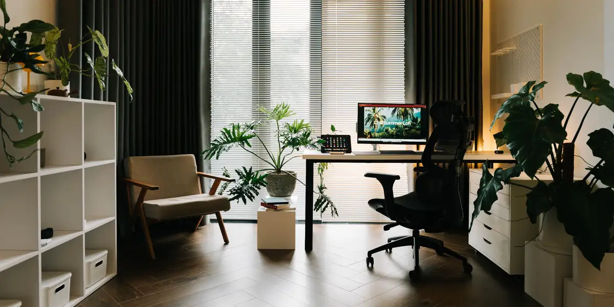 Black ergonomic mesh chair with a headrest at a white desk in a plant-filled home office with tall windows