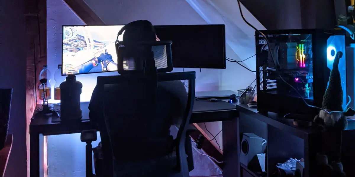 Gamer in a mesh office chair with a headrest at a dual-monitor PC setup in a dimly lit room
