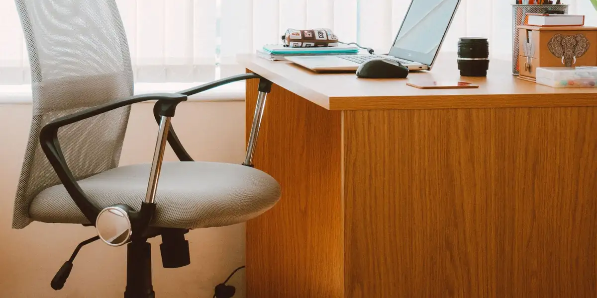 Grey mesh-back swivel chair beside a wooden desk with a laptop and window blinds