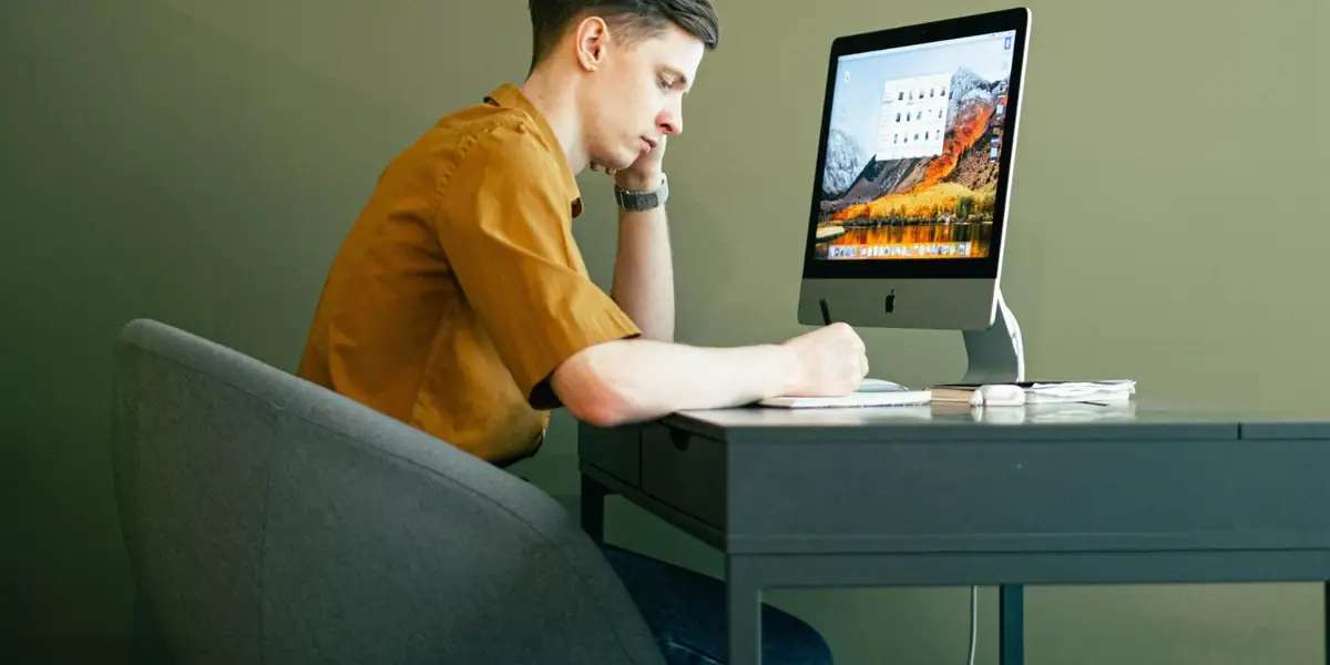 Man leaning forward and writing at a desk with a desktop computer while seated in a tub chair