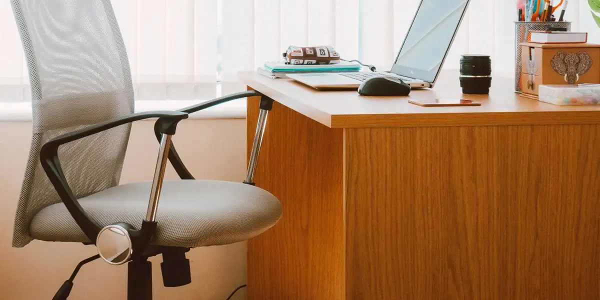 Gray mesh-back office chair beside a wooden desk with a laptop in front of bright window blinds