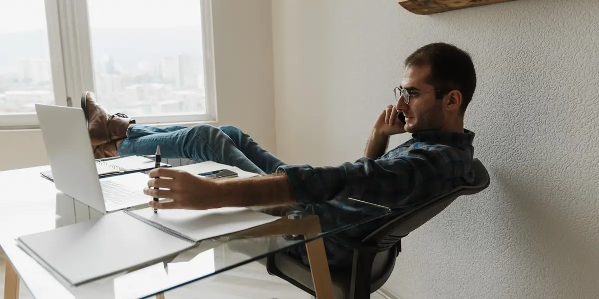 Man reclining in a mesh office chair with his feet on the desk while taking a phone call