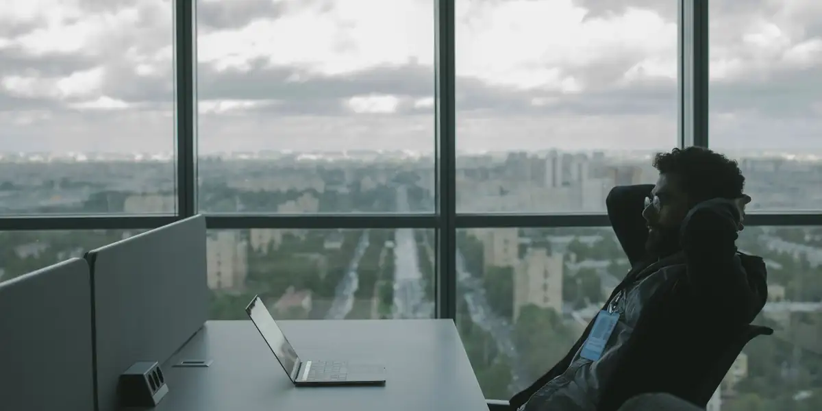 Man reclining in an office chair with his hands behind his head beside a city-view window