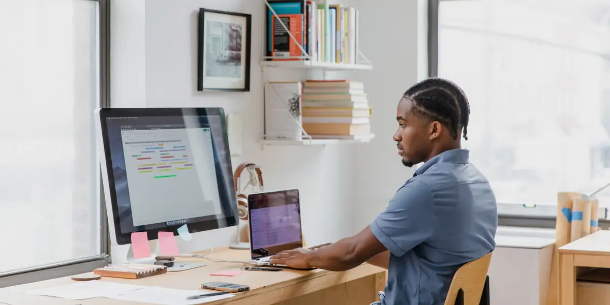 Man sitting upright typing on a laptop beside a large monitor at a wooden desk by a window